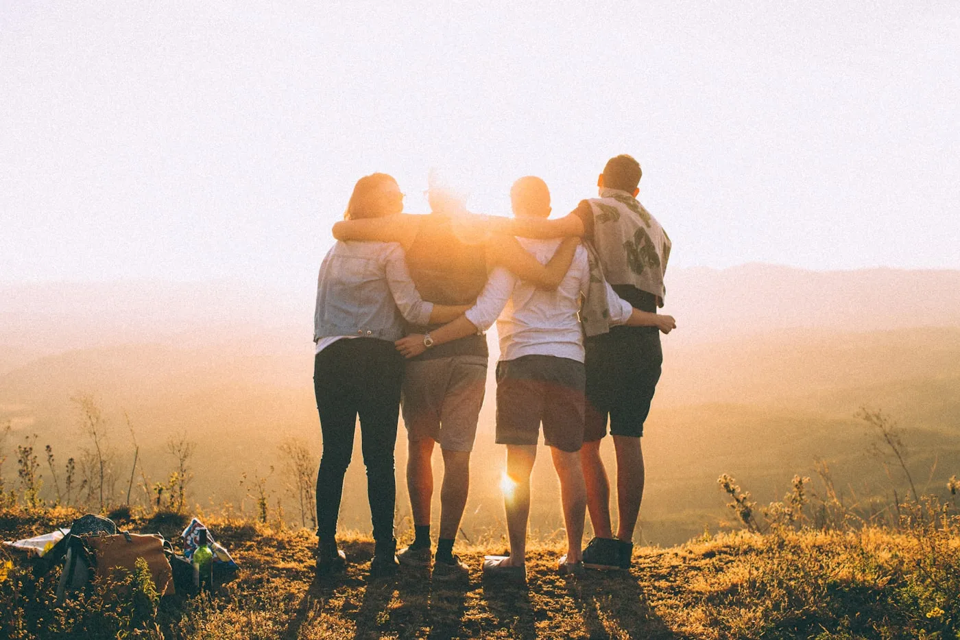 Five friends standing shoulder to shoulder with arms around each other, watching the sun set.