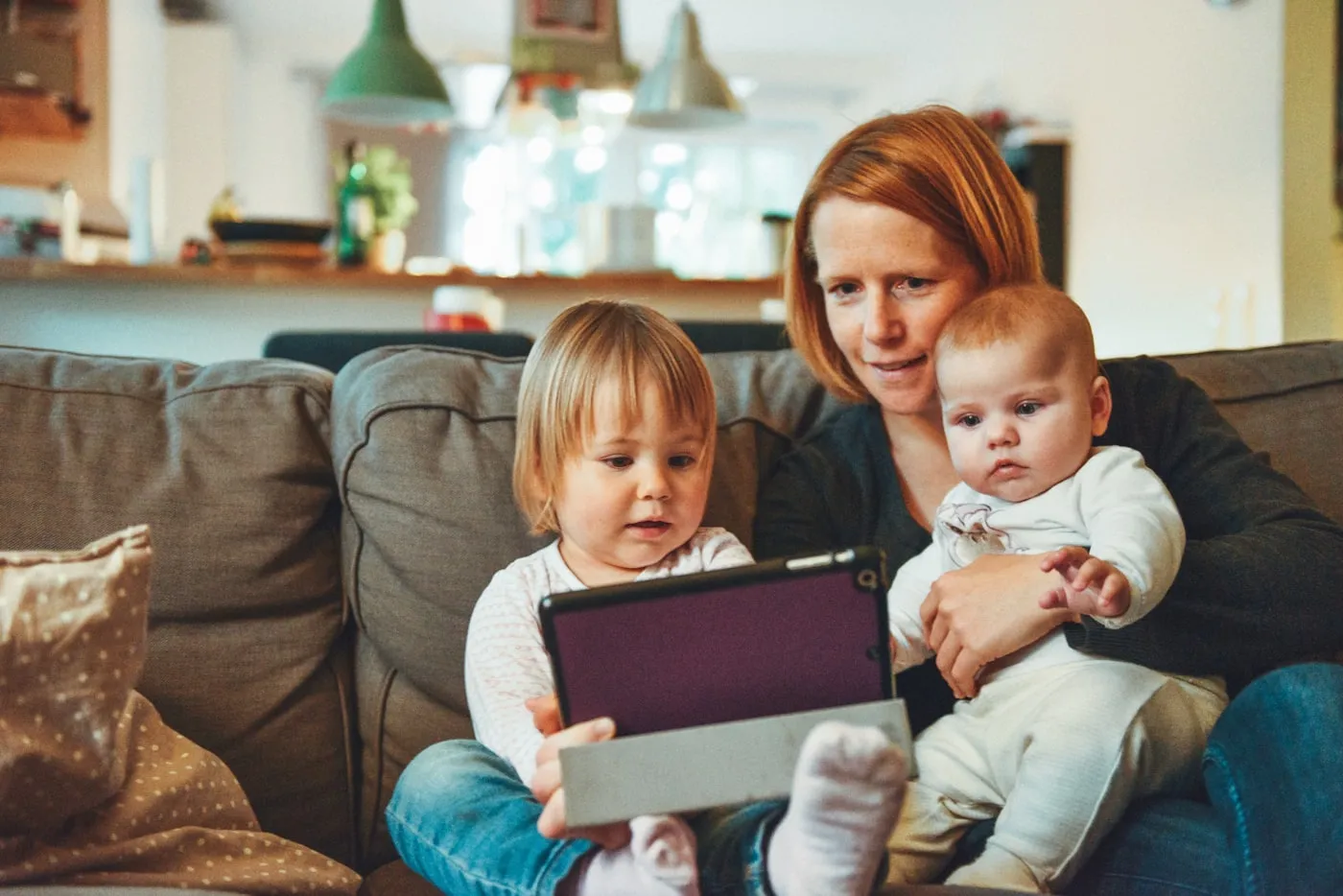 A parent on a couch with a toddler and a baby, looking at a picture book together.